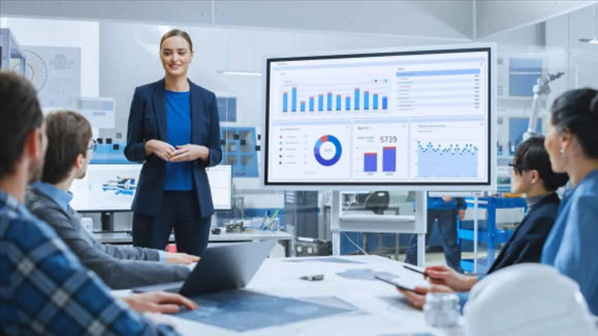 Mujer profesional presenta análisis de datos en pantalla grande a un equipo en reunión. Gráficos y cuadros de mando visibles. Entorno de oficina.