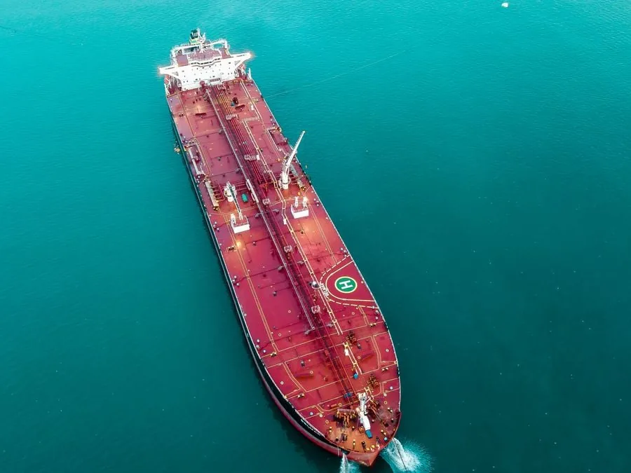Vista aérea de un gran superpetrolero de color rojo oscuro navegando en aguas turquesas, mostrando su cubierta con tuberías y una helisuperficie.