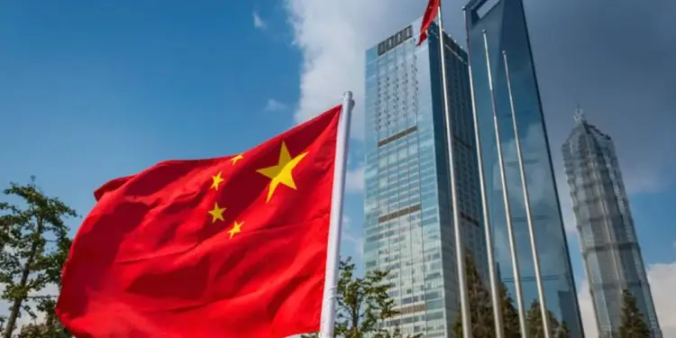 Bandera de China ondeando frente a un grupo de rascacielos modernos de cristal bajo un cielo azul. Símbolo de economía y negocios.