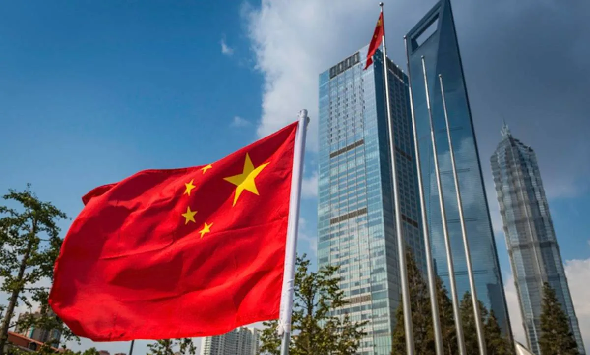 Bandera de China ondeando frente a un grupo de rascacielos modernos de cristal bajo un cielo azul. Símbolo de economía y negocios.
