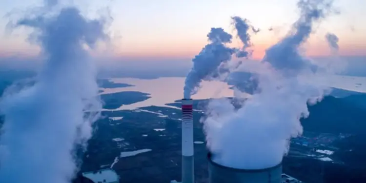 Vista aérea de una planta de energía con grandes chimeneas emitiendo humo/vapor denso al atardecer, sobre un paisaje con lagos y vegetación.