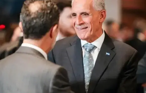 Dos hombres de negocios en trajes formales interactúan en un evento. Uno sonríe, lleva un pin con franjas azul y blanca en su solapa.