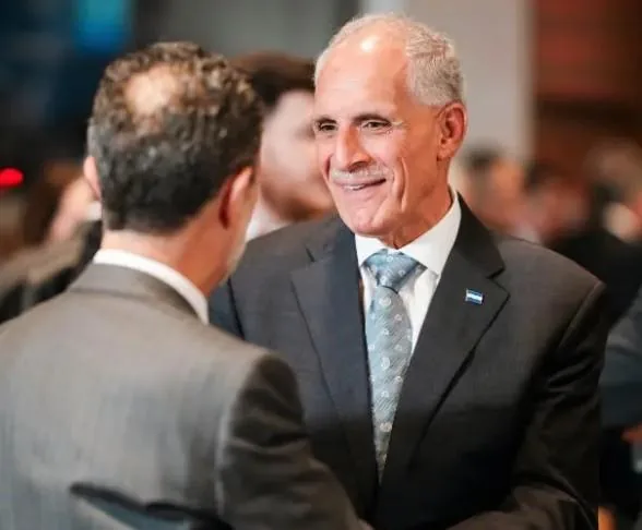 Dos hombres de negocios en trajes formales interactúan en un evento. Uno sonríe, lleva un pin con franjas azul y blanca en su solapa.
