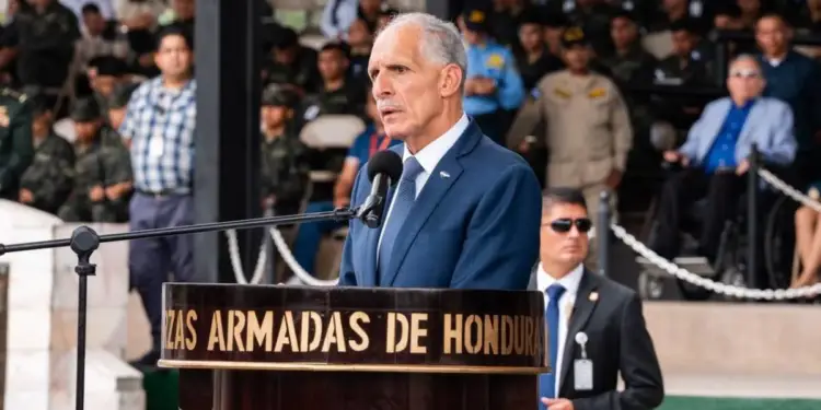 Hombre en traje azul habla en podio con micrófono, inscripción 'Fuerzas Armadas de Honduras'. Guardia de seguridad y público diverso.