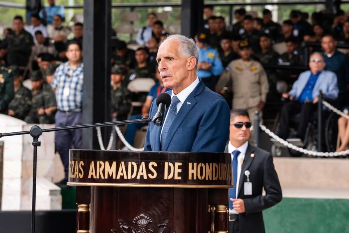 Hombre en traje azul habla en podio con micrófono, inscripción 'Fuerzas Armadas de Honduras'. Guardia de seguridad y público diverso.