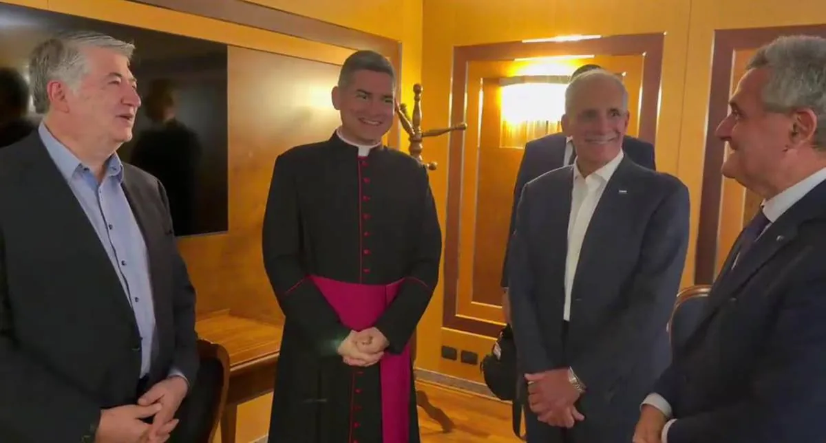 Cuatro hombres, uno vestido de clérigo y tres con trajes de negocios, sonríen y conversan en un entorno profesional interior.