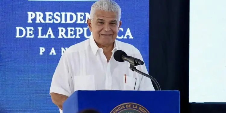 Hombre maduro con cabello gris vistiendo camisa blanca hablando frente a un micrófono en un podio con sello oficial.