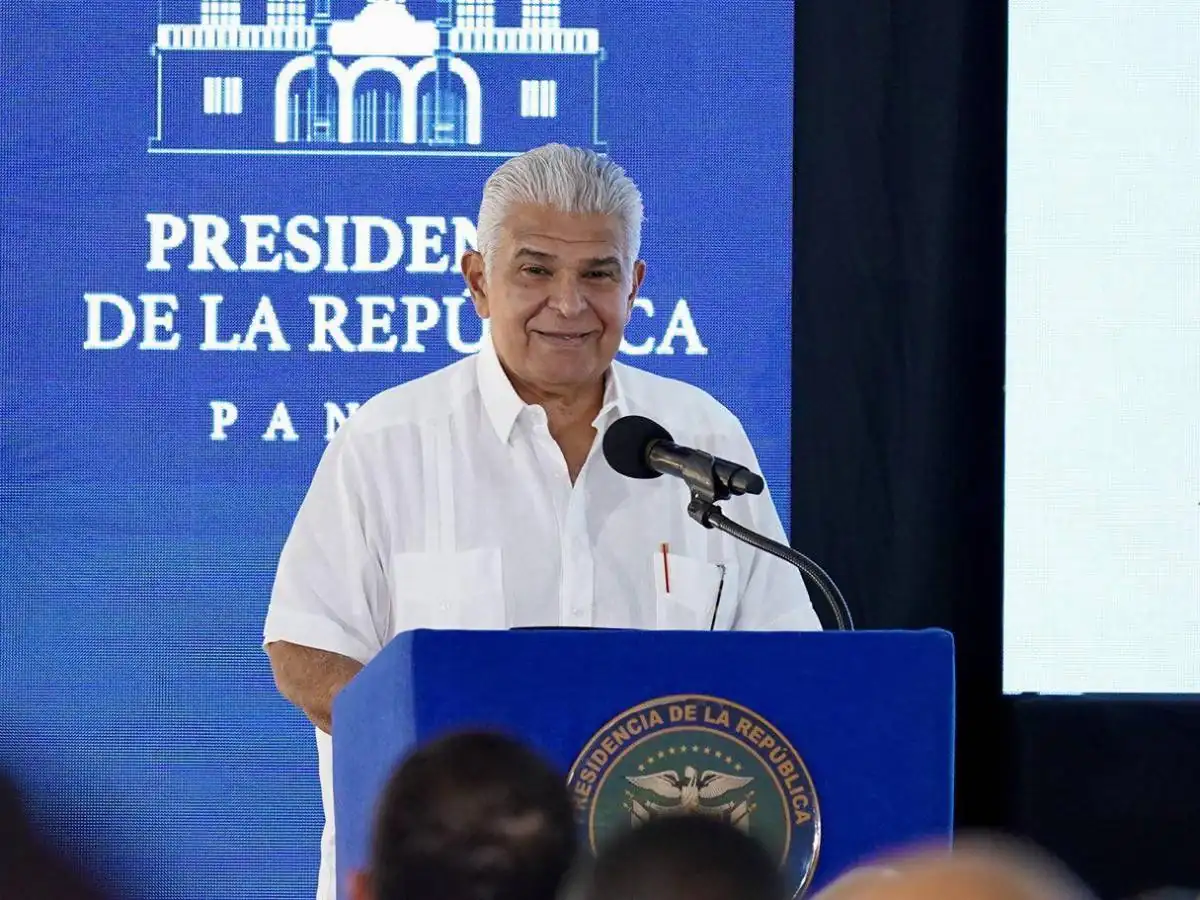 Hombre maduro con cabello gris vistiendo camisa blanca hablando frente a un micrófono en un podio con sello oficial.