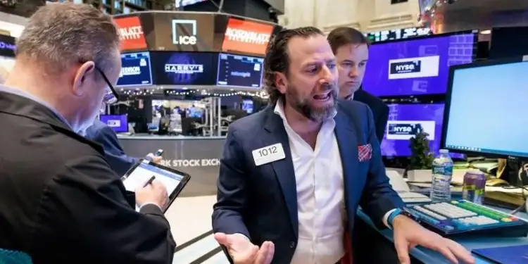 Operadores bursátiles en un piso de negociación, uno gesticula mientras otro usa una tablet. Pantallas con datos financieros y el logo NYSE.
