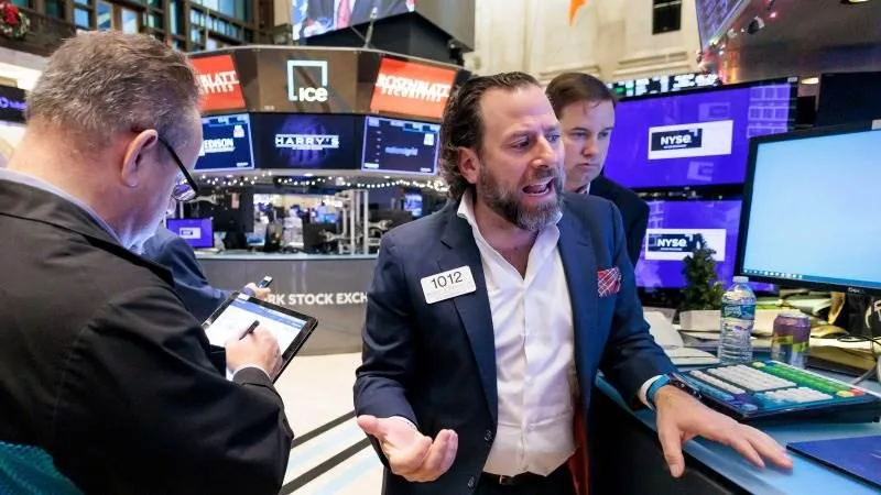 Operadores bursátiles en un piso de negociación, uno gesticula mientras otro usa una tablet. Pantallas con datos financieros y el logo NYSE.