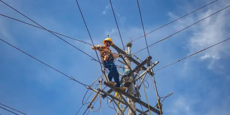 Un trabajador con casco amarillo y arnés de seguridad trabaja en un poste de electricidad, revisando cables y conexiones contra un cielo azul con nubes dispersas.