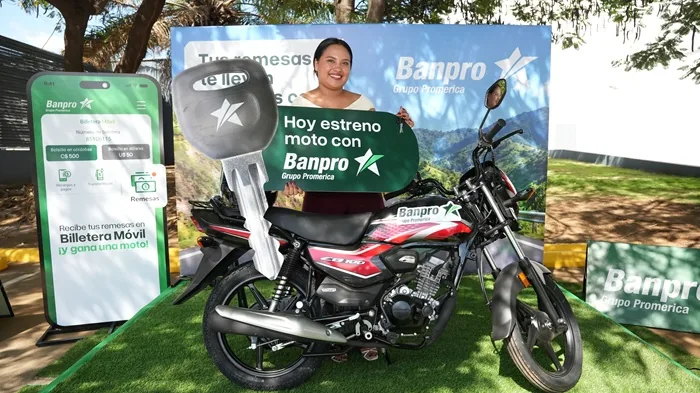 Mujer sonriente sosteniendo una llave gigante junto a una motocicleta y carteles promocionales de Banpro Grupo Promerica.
