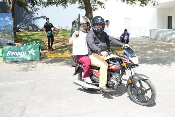 Hombre y mujer sonríen en motocicleta Honda negra y roja, ambos con cascos, frente a banners de Banpro Grupo Promerica.