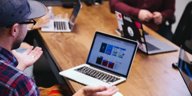 Personas en una reunión de negocios alrededor de una mesa de madera, trabajando en laptops que muestran gráficos y datos. Uno de ellos gesticula con las manos.