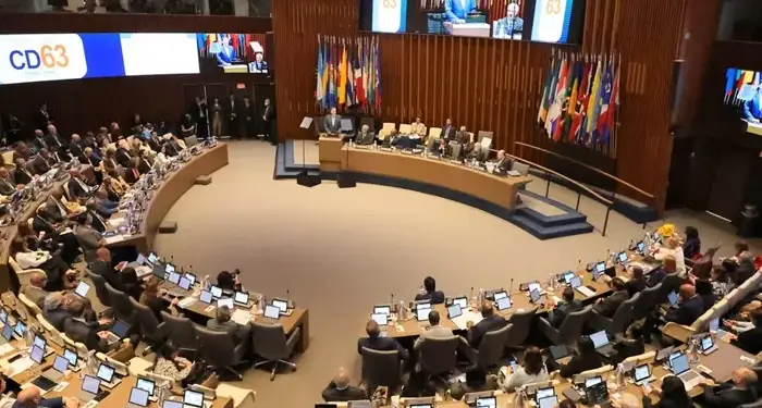 Gran sala de conferencias con delegados en escritorios curvos usando laptops. Un orador en podio, panel de expertos, banderas y pantalla 'CD63' al fondo.