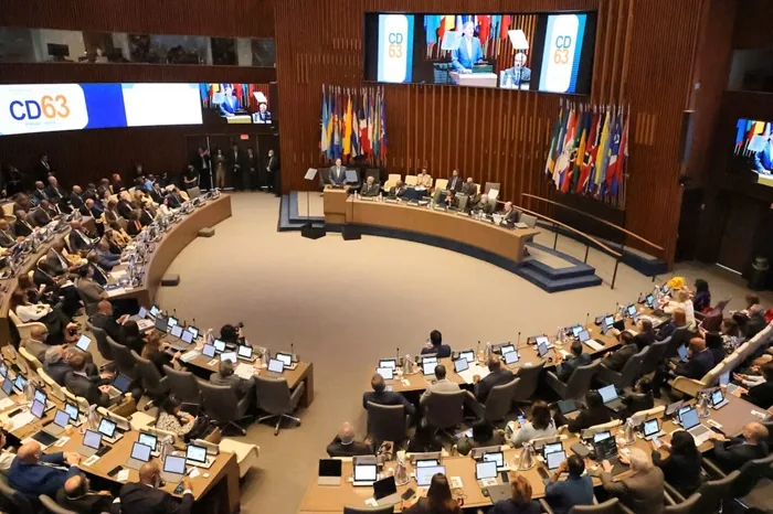 Gran sala de conferencias con delegados en escritorios curvos usando laptops. Un orador en podio, panel de expertos, banderas y pantalla 'CD63' al fondo.