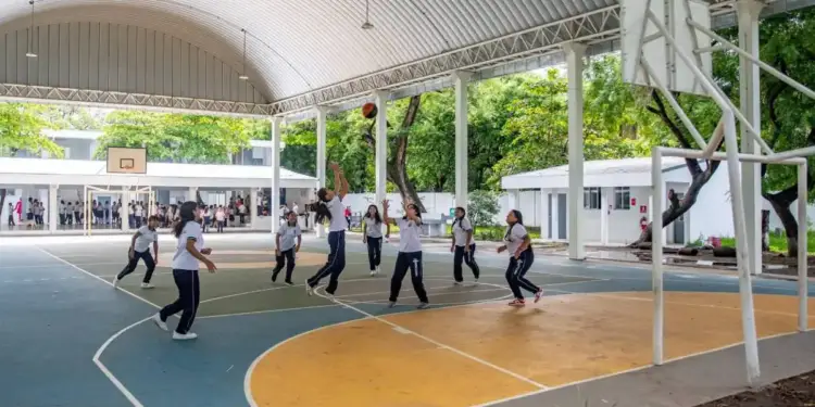 Grupo de jóvenes estudiantes con uniforme practicando baloncesto en una cancha deportiva techada al aire libre.