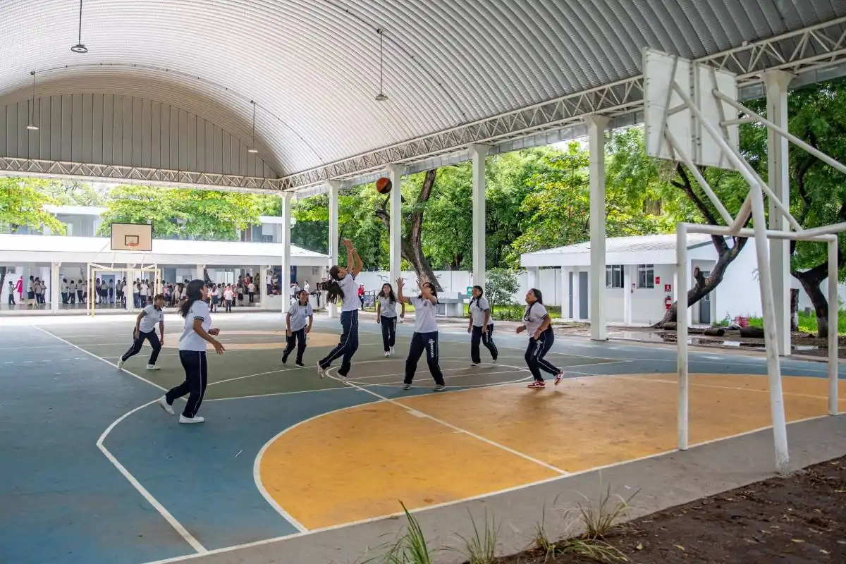 Grupo de jóvenes estudiantes con uniforme practicando baloncesto en una cancha deportiva techada al aire libre.