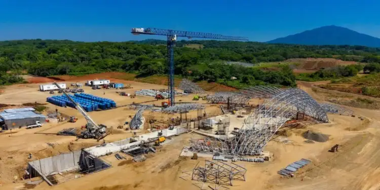 Vista aérea de un sitio de construcción a gran escala con grúas, maquinaria pesada, estructuras metálicas y materiales apilados bajo un cielo azul claro.