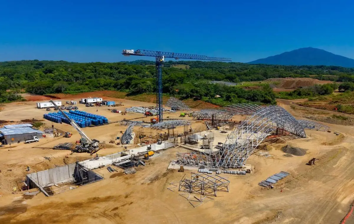 Vista aérea de un sitio de construcción a gran escala con grúas, maquinaria pesada, estructuras metálicas y materiales apilados bajo un cielo azul claro.