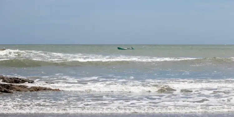 Pequeño bote navegando en el mar con olas rompiendo cerca de la orilla rocosa bajo un cielo despejado.
