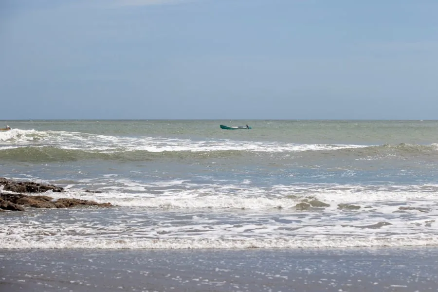 Pequeño bote navegando en el mar con olas rompiendo cerca de la orilla rocosa bajo un cielo despejado.
