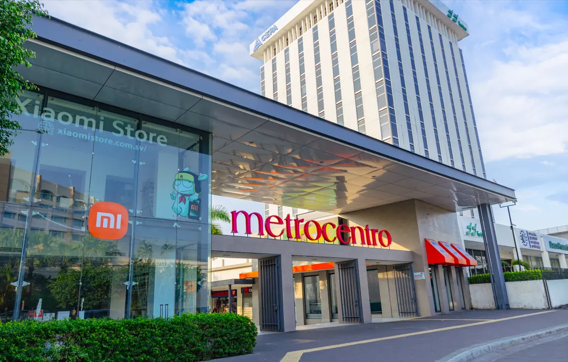 Fachada de un complejo comercial con la tienda Xiaomi, el letrero "metrocentro" y un edificio corporativo alto. Se observan logos de CEPA y Banco Promerica.