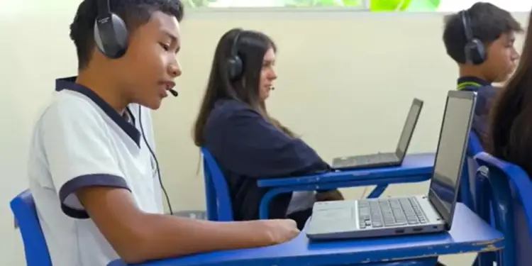 Jóvenes estudiantes con auriculares y micrófonos utilizando computadoras portátiles en un salón de clases.