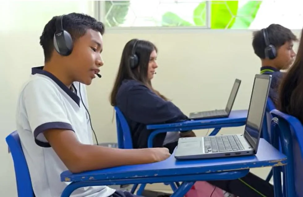 Jóvenes estudiantes con auriculares y micrófonos utilizando computadoras portátiles en un salón de clases.