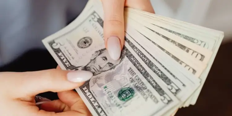 Primer plano de manos sosteniendo y contando un fajo de billetes de cinco dólares estadounidenses, con el retrato de Abraham Lincoln visible.