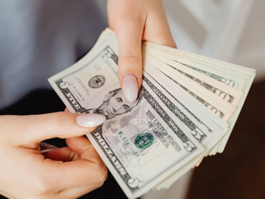 Primer plano de manos sosteniendo y contando un fajo de billetes de cinco dólares estadounidenses, con el retrato de Abraham Lincoln visible.