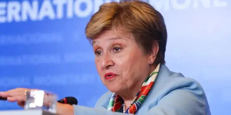 Primer plano de una mujer de mediana edad con blazer azul y pañuelo colorido, hablando en un podio con micrófono. Fondo azul con texto 'INTERNATIONAL MONETARY'.