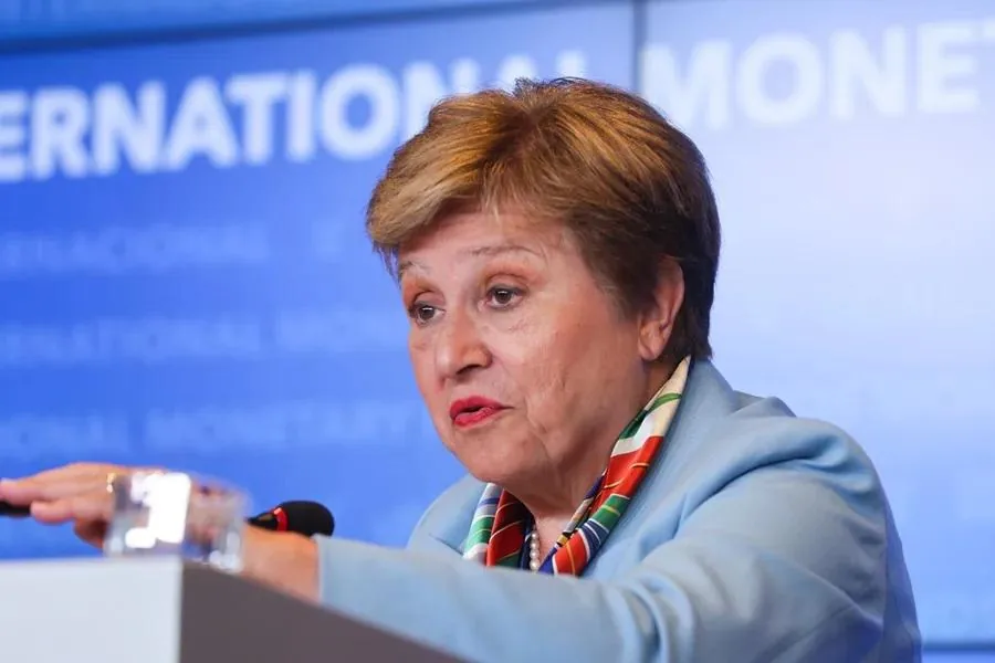 Primer plano de una mujer de mediana edad con blazer azul y pañuelo colorido, hablando en un podio con micrófono. Fondo azul con texto 'INTERNATIONAL MONETARY'.