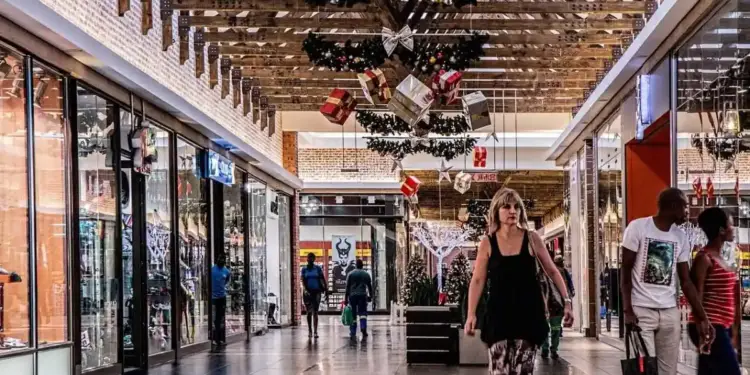 Personas caminando por el pasillo de un centro comercial decorado con adornos navideños y tiendas de retail.