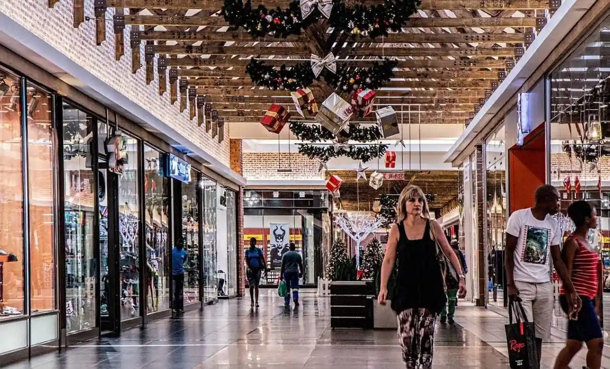 Personas caminando por el pasillo de un centro comercial decorado con adornos navideños y tiendas de retail.