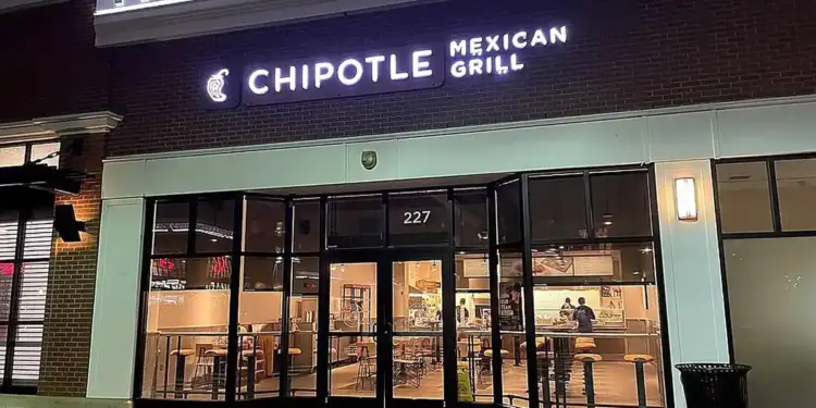 Fachada nocturna de un restaurante Chipotle Mexican Grill con letrero luminoso, grandes ventanales y clientes en el interior.