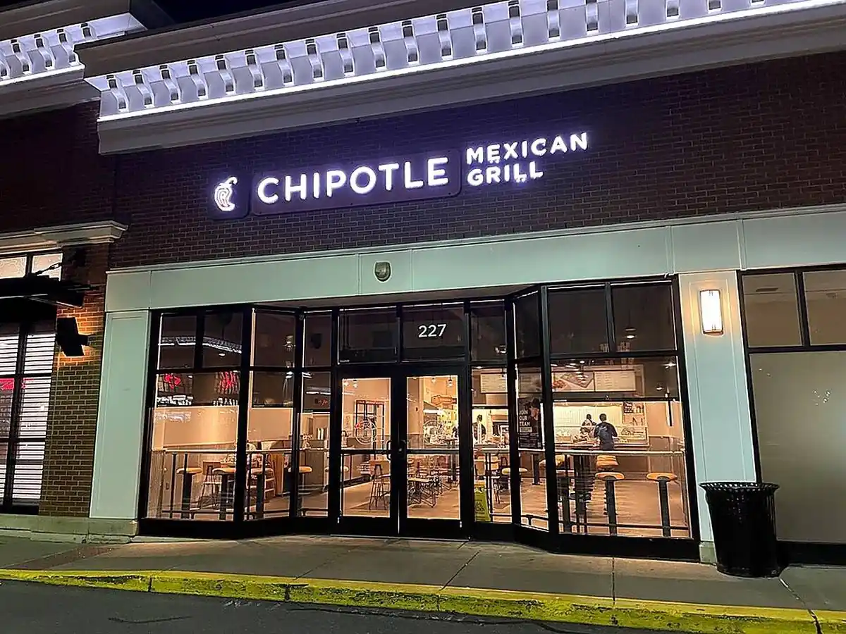 Fachada nocturna de un restaurante Chipotle Mexican Grill con letrero luminoso, grandes ventanales y clientes en el interior.