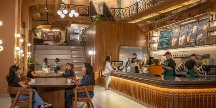 Amplia cafetería de diseño contemporáneo con detalles en madera. Personas interactúan en mesas y área de descanso escalonada, personal atiende.