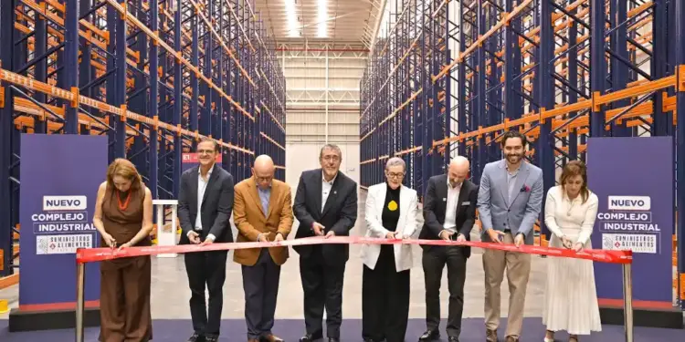 Ocho profesionales cortan una cinta roja en la inauguración de un nuevo complejo industrial con estanterías de almacén vacías. Fondo azul con texto 'Nuevo Complejo Industrial Suministros Alimentos'.