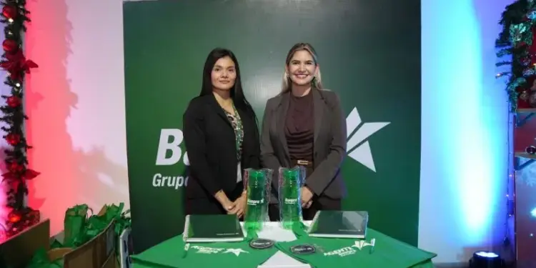 Dos mujeres profesionales posando frente a un panel verde con logotipos corporativos y una mesa con material promocional.