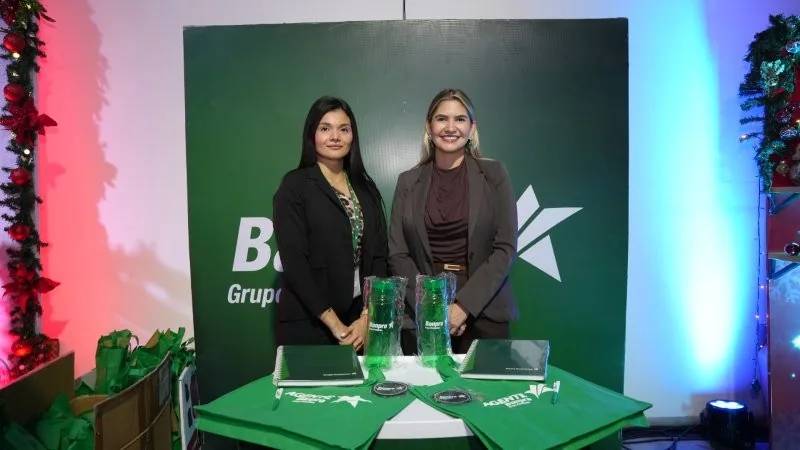 Dos mujeres profesionales posando frente a un panel verde con logotipos corporativos y una mesa con material promocional.