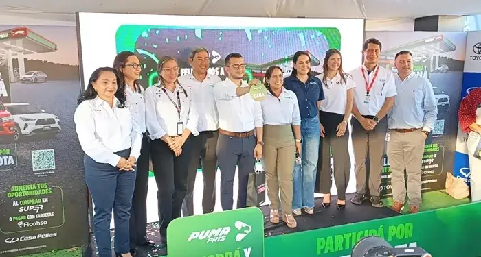 Grupo de diez profesionales en escenario verde. Hombre central sostiene objeto 'GANA'. Banners de gasolinera, autos y servicios financieros.