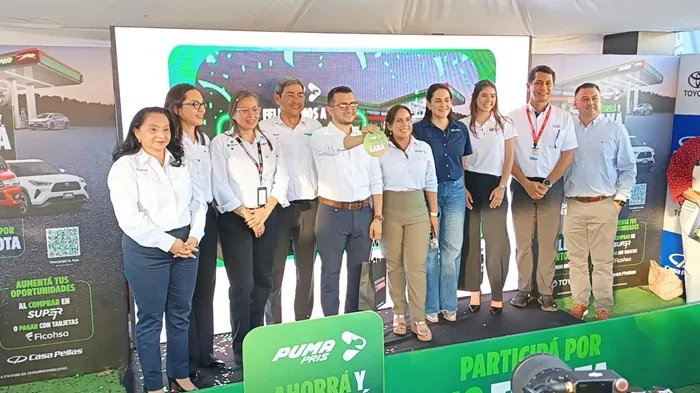 Grupo de diez profesionales en escenario verde. Hombre central sostiene objeto 'GANA'. Banners de gasolinera, autos y servicios financieros.