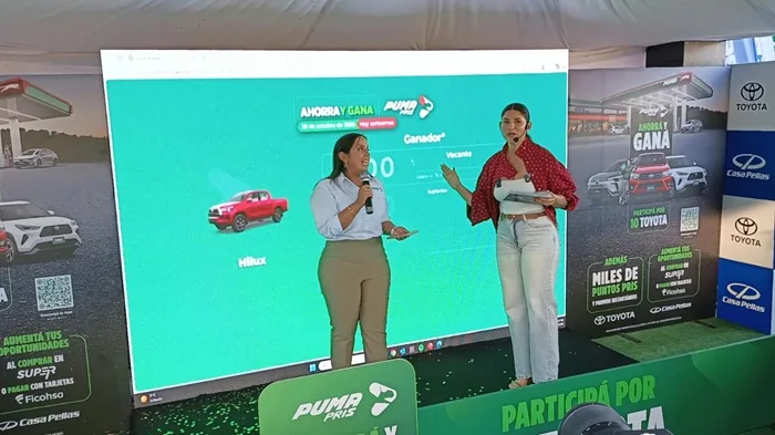 Dos mujeres presentan una pantalla digital que muestra la promoción 'Ahorra y Gana' con un vehículo Toyota Hilux y detalles de premios, junto a un cartel publicitario de la misma promoción.