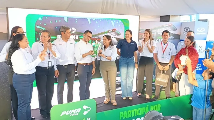 Un grupo de personas vestidas formalmente, hombres y mujeres, posan en un escenario frente a una pantalla grande que muestra una estación de servicio. Algunos aplauden mientras otros sostienen premios o bolsas.