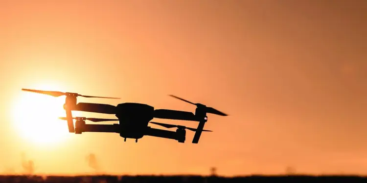Silueta de un dron moderno volando contra un cielo anaranjado brillante al atardecer, con el sol visible.