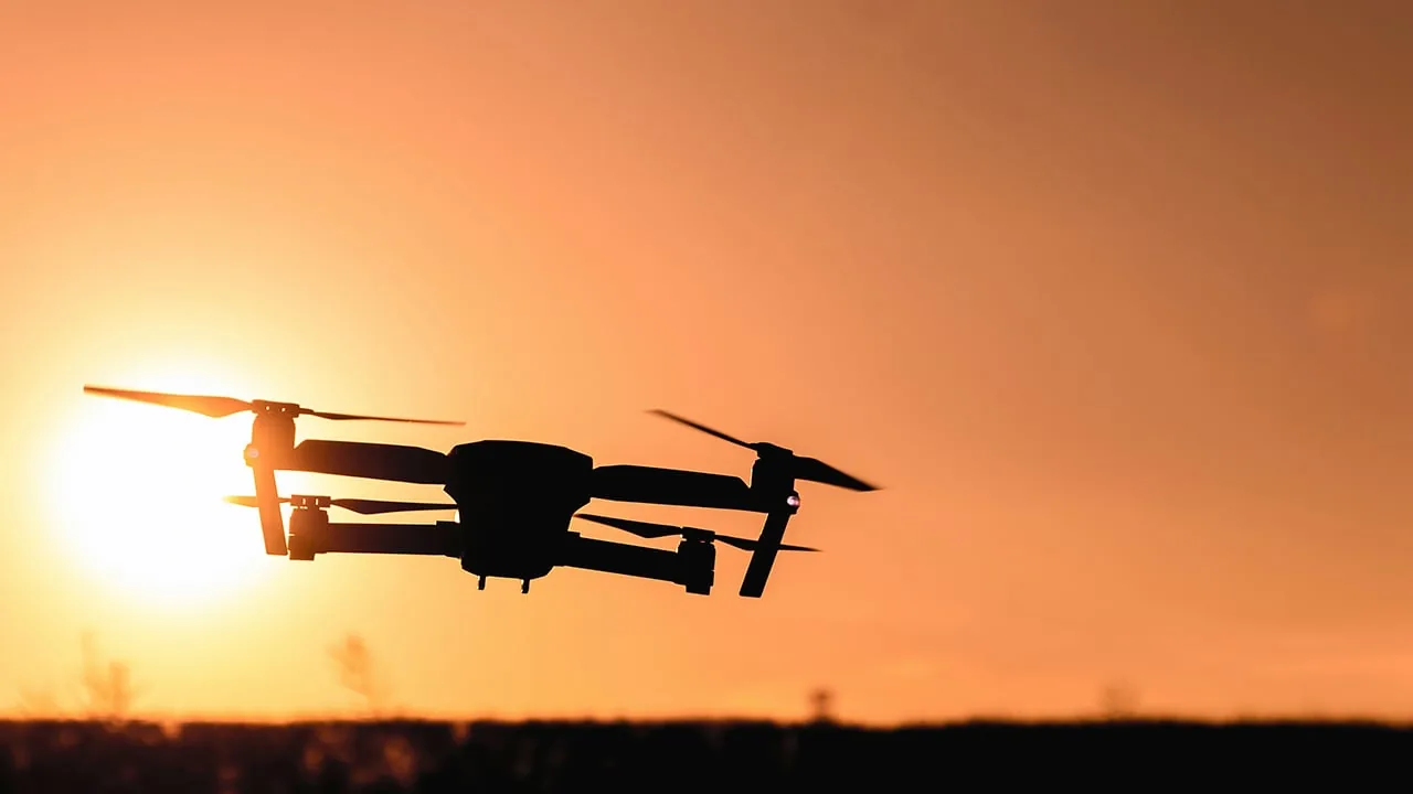 Silueta de un dron moderno volando contra un cielo anaranjado brillante al atardecer, con el sol visible.