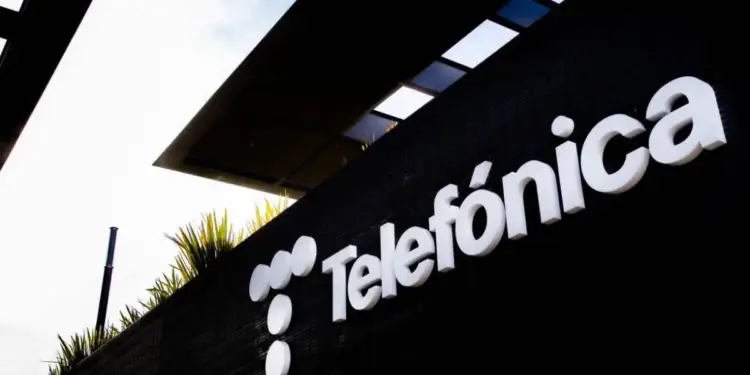 Primer plano del letrero blanco 3D de 'Telefónica' y su logo de tres círculos sobre una pared negra texturizada. Fondo con estructura moderna y vegetación.