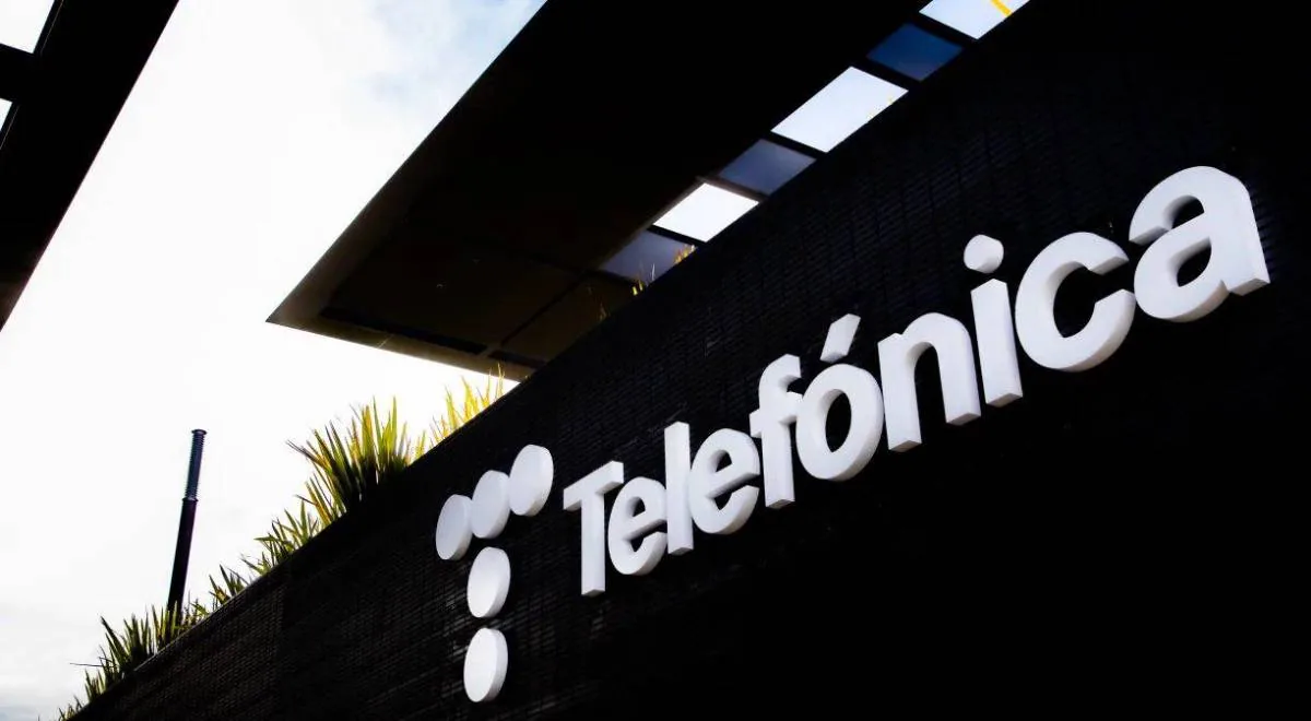 Primer plano del letrero blanco 3D de 'Telefónica' y su logo de tres círculos sobre una pared negra texturizada. Fondo con estructura moderna y vegetación.