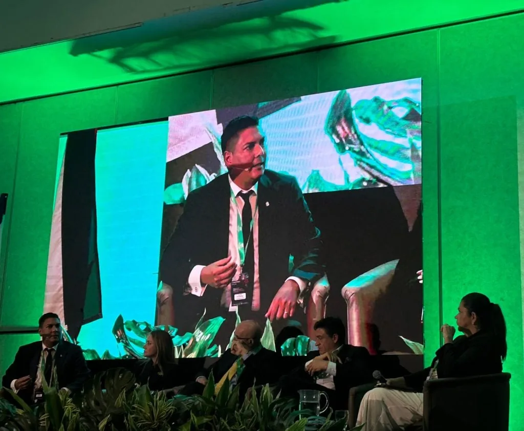 Panelistas en una conferencia de negocios frente a una gran pantalla que muestra a uno de los ponentes.